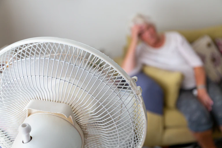 Blurred image of a woman with gray hair leaning on her elbow on a sofa with an electric fan in the foreground
