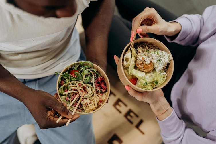 Two people eat noodles from takeaway bowls.