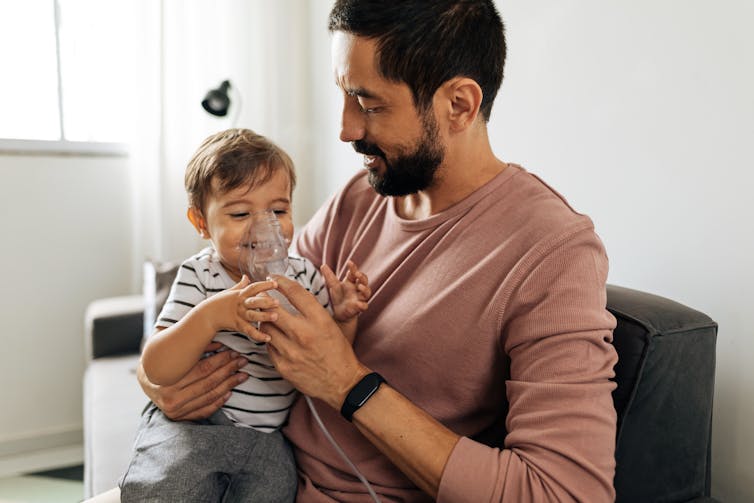 A father helps a small smiling boy to breathe through a nebuliser.