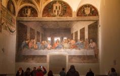 A mural depicting Jesus Christ surrounded by the apostles at the Last Supper, painted on a wall in front of several visitors.