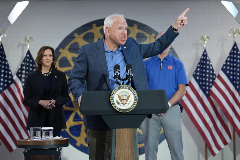 A man in a blue jacket and blue shirt speaking into microphones and gesturing with his left hand.