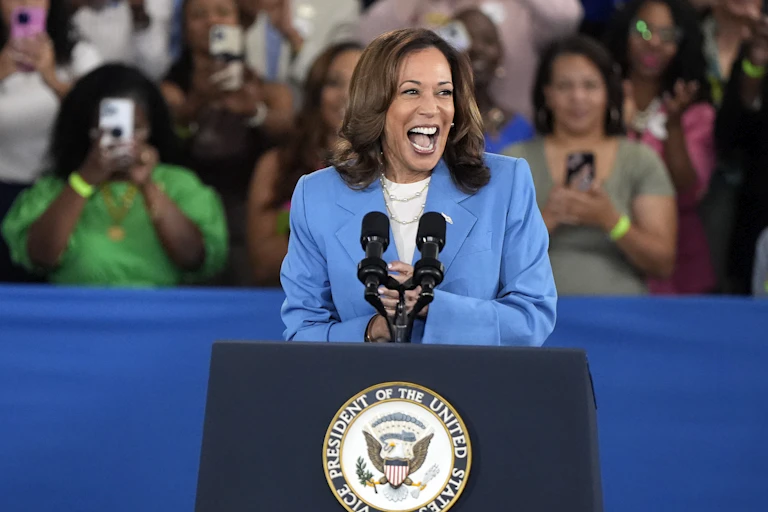 A dark-haired woman in a blue suit laughs behind a lectern at a campaign rally; women behind her are seen taking photos with the phones.