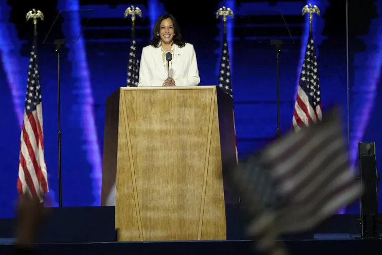 A woman in a cream suit smiles from behind a lectern as people in the crowd wave American flags.