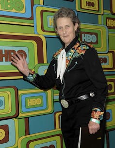 Temple Grandin, a person in a black suit with a colourful shirt underneath, in front of a wall emblazoned with 'HBO'.
