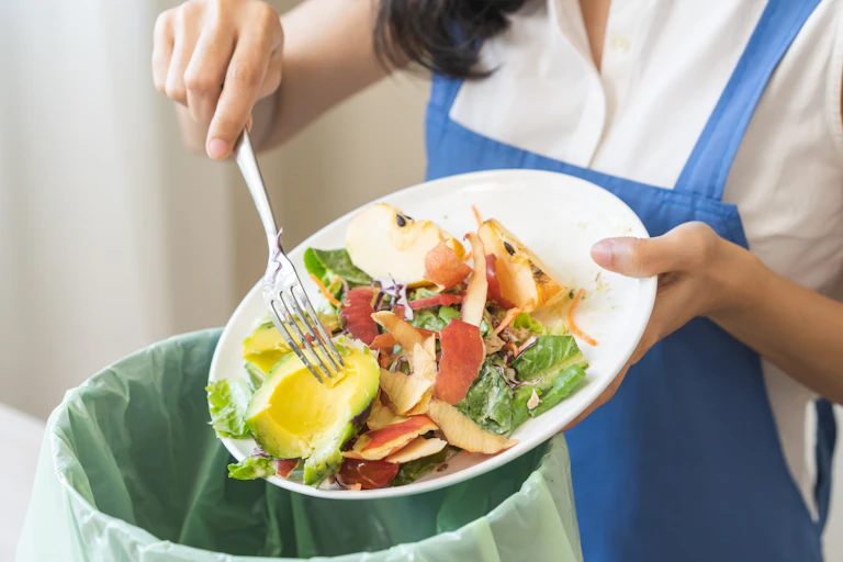 woman in blue and white clothes scraping food veg scraps from white bowl into compost bin