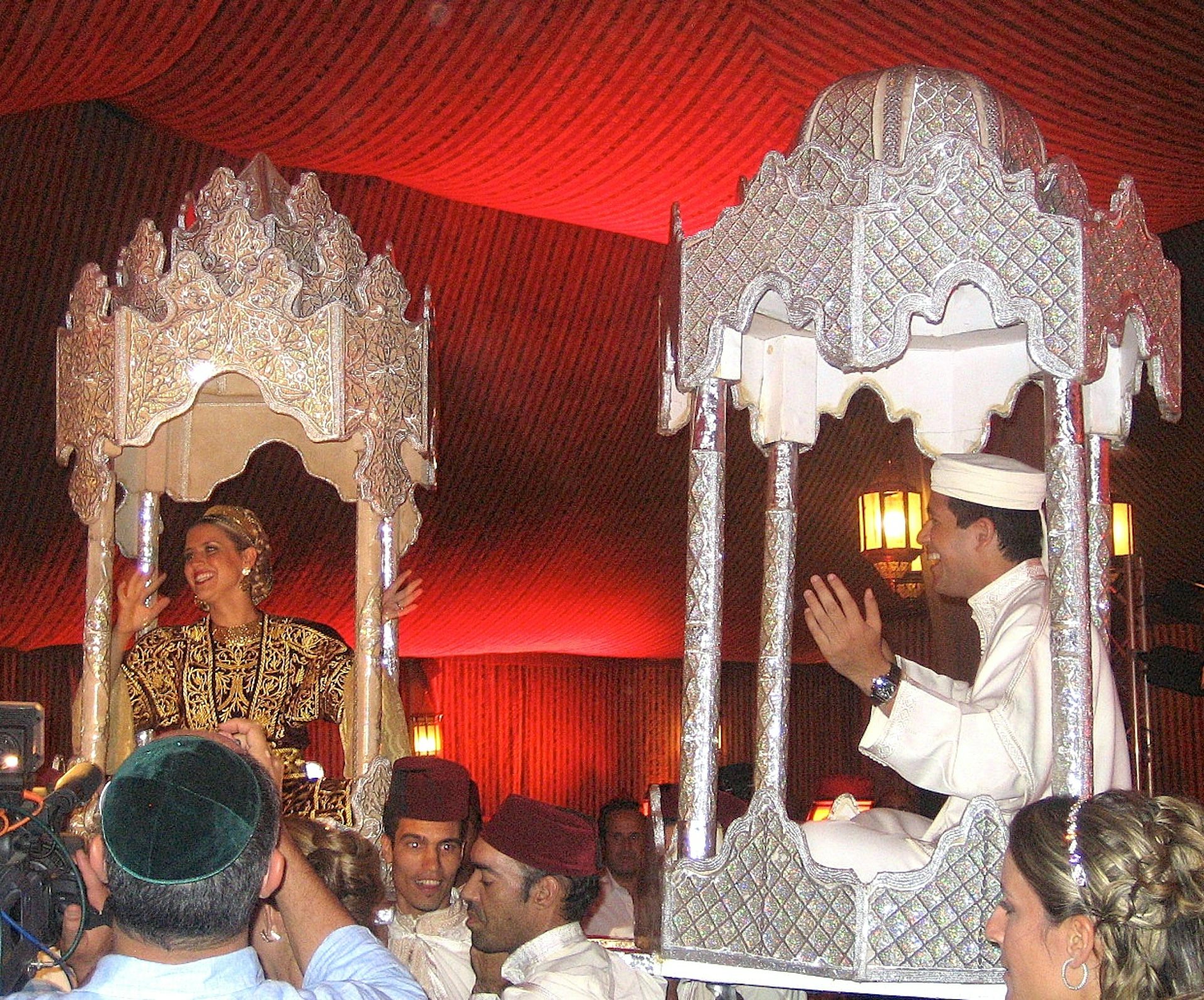 A man and woman each sit in an ornate, canopied chair carried by several men, inside a red rom.