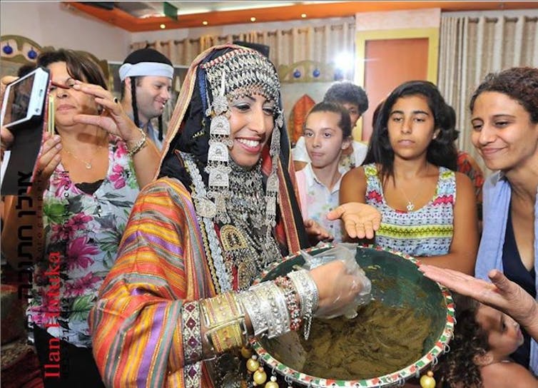 A woman in an ornate, colorful robe with jewelry and a headdress scoops a paste out of a bowl as young women hold out their hands.