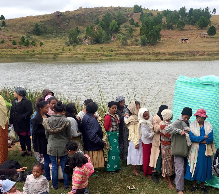A line of women wait at the banks of a wide river, with a curtain blocking off part of the view.
