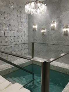 A small room with white marble walls, ornate lighs, and a small pool of teal-colored water with steps leading down into it.