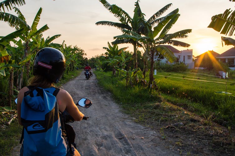 A woman on a scooter on a rural road in the tropics at dusk.