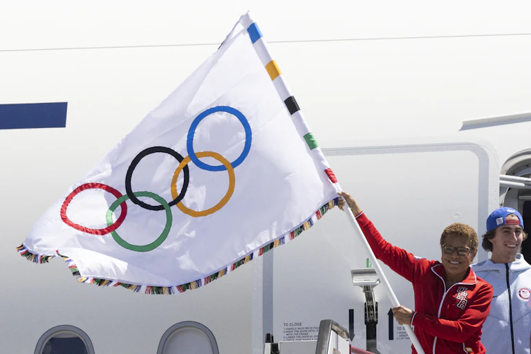 A woman in a red workout jacket stands outside a jet plane holding the Olympic flag