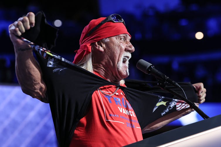 An older white man with a white mustache wears a red bandana and a red shirt that says 'Trump/Vance.'