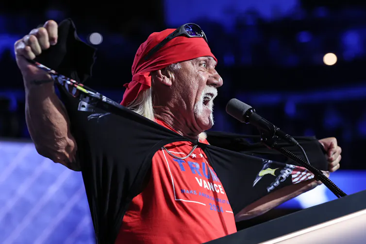 An older white man with a white mustache wears a red bandana and a red shirt that says 'Trump/Vance.'