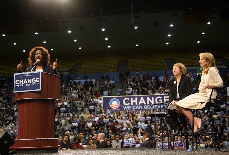 A woman stands at a podium that has the sign 'Change we can believe in' in front of a large crowd of people.