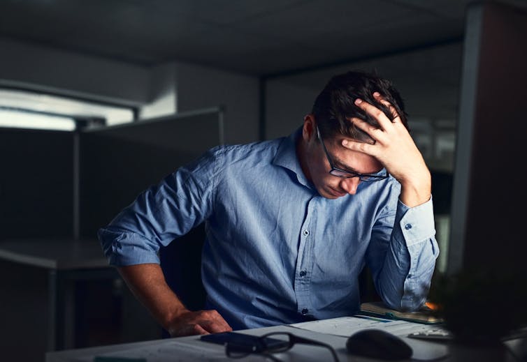 A young man sitting in a dark office rests his head against his hand with an exhausted look on his face