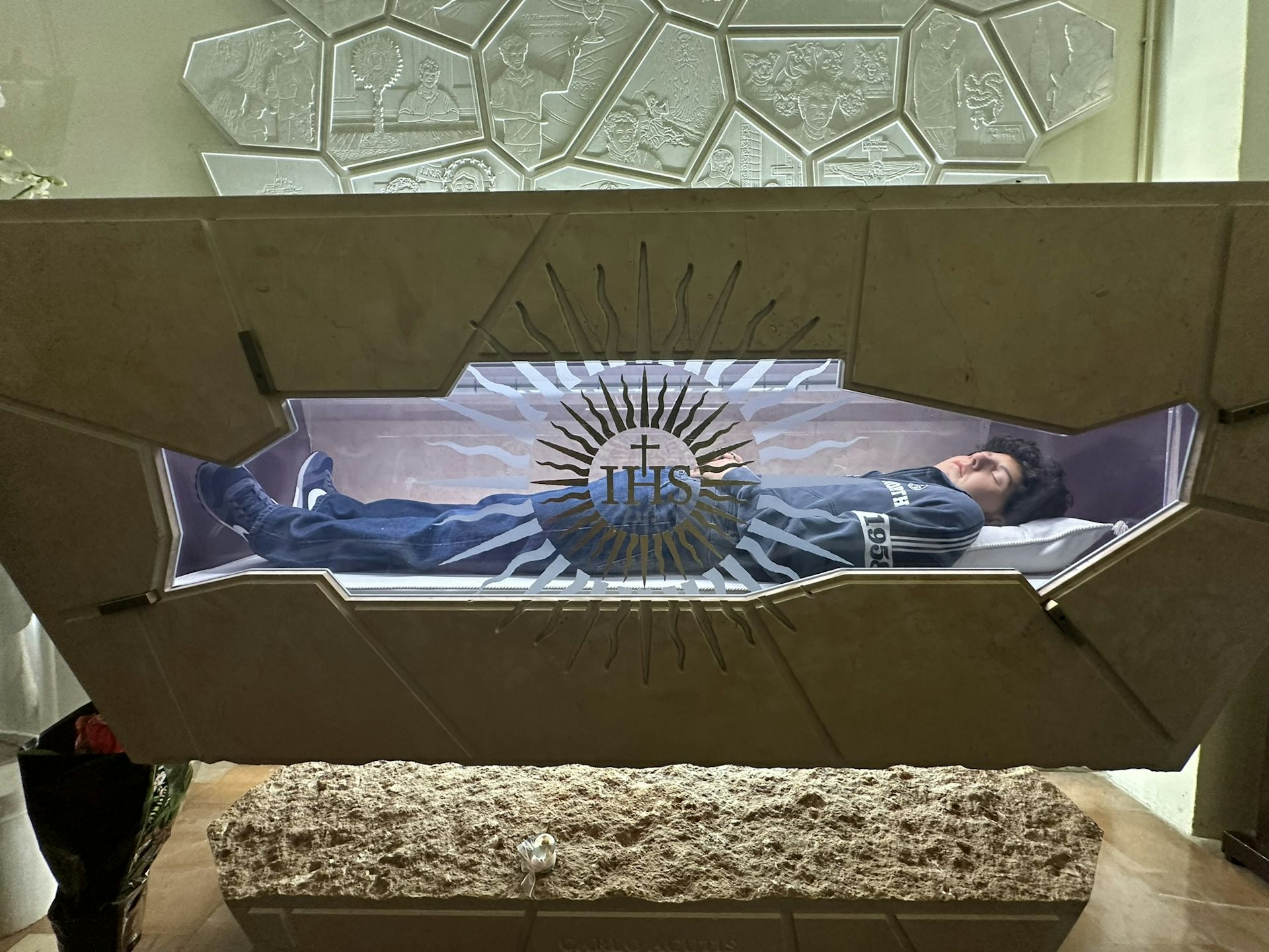 The body of a young boy with black, curly hair, dressed in jeans and sneakers, seen through a glass-covered opening in a sarcophagus.