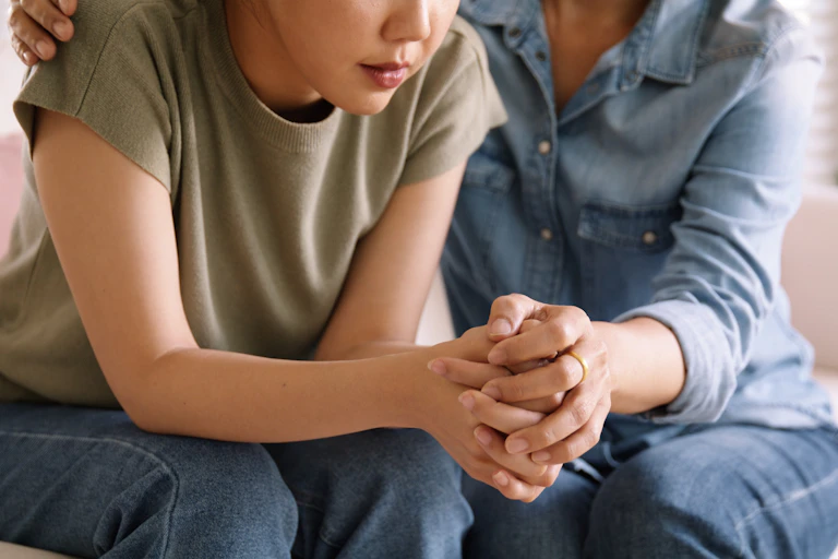 A woman sits beside another woman, both only seen from the shoulders down, and holds her hand and has an arm around her shoulders