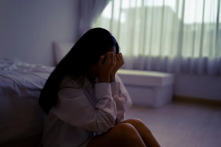 A young woman sits on the ground of a bedroom with her head buried in her hands