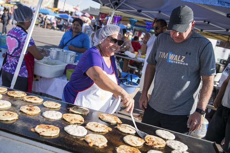 A man in a t-shirt and baseball cap next to a woman frying dough on a large grill outside.