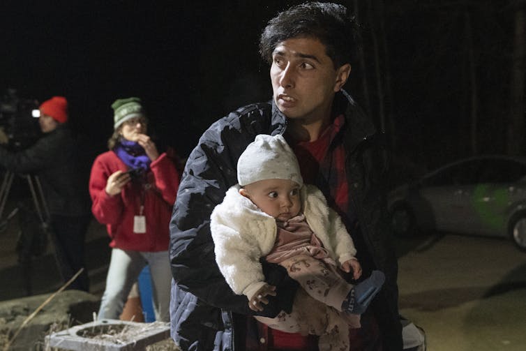 A man with a fearful look on his face carries a small baby on a darkened road.