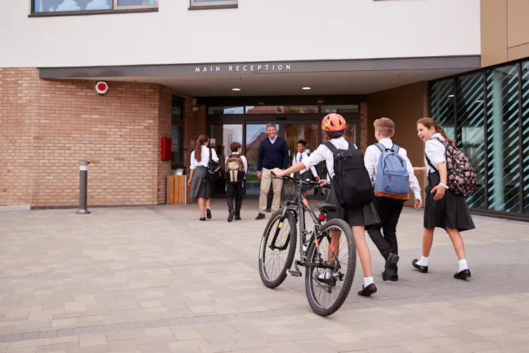 A group of students walk to the main entrance of a high school. A male teacher stands at the entrance, smiling.