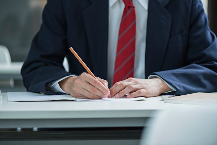 A teenage boy in a blazer and tie writes on a sheet of paper on a desk.