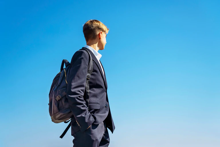 A teenage boy in a dark blazer and backpack looks over his shoulder against a bright blue sky.