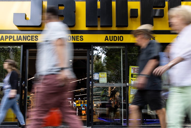 Blurred customers walk in front of a JB Hi-Fi store