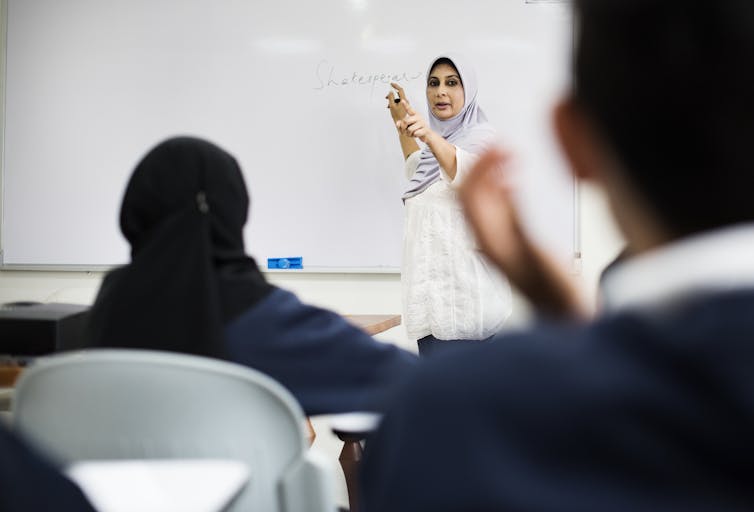 A teacher points to a student as she writes on the whiteboard.