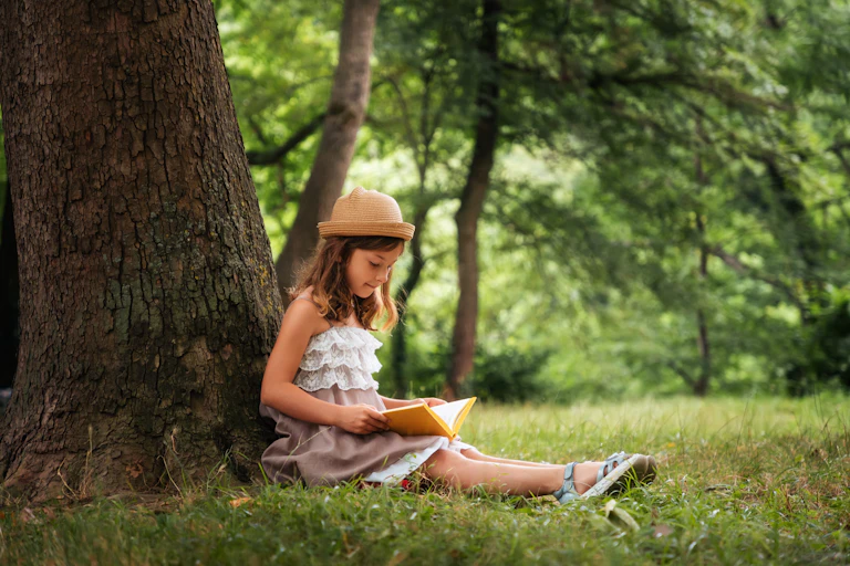 A school-aged girl in a straw hat and dress is sitting on the grass by a tree and reading a book.