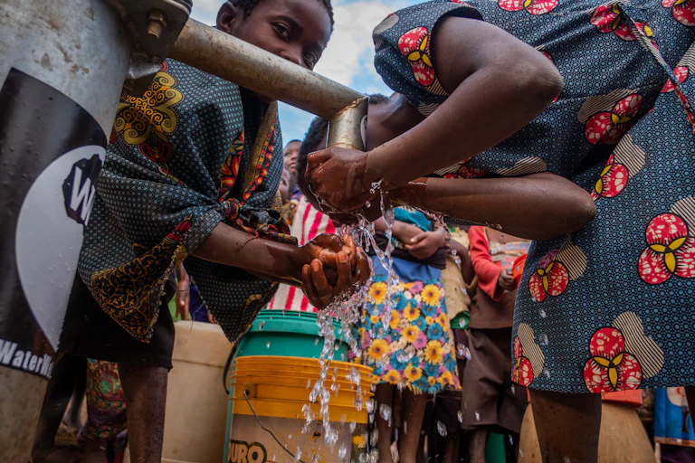 A group of young people gather around a standpipe, drinking the water coming from it or using it to wash their hands