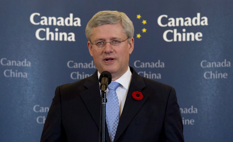 A grey-haired man wearing glasses speaks into a microphone in front of a banner that reads Canada China.
