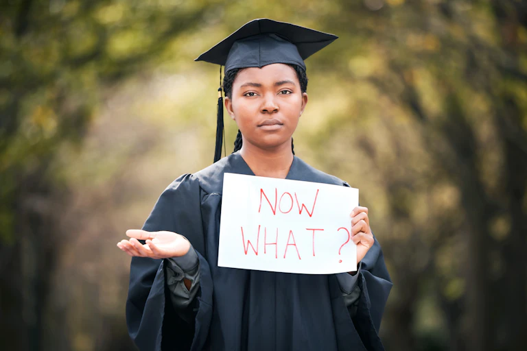 Young person wearing an academic gown and mortarboard, holding a sign that says 'now what?'