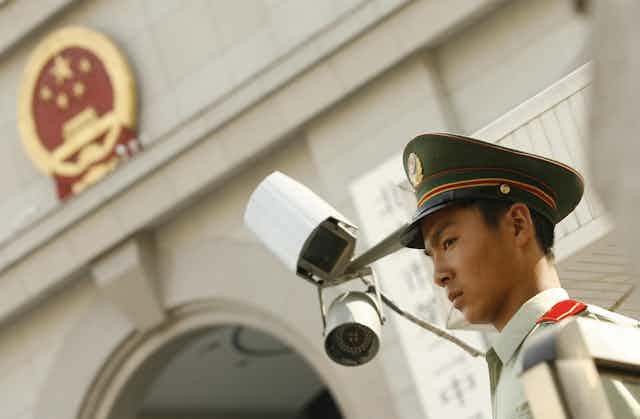A man in a uniform with a stern expression stands in front of a pair of surveillance cameras on the outside of a concrete building