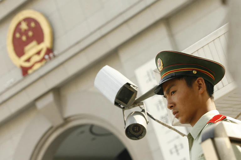 A man in a uniform with a stern expression stands in front of a pair of surveillance cameras on the outside of a concrete building
