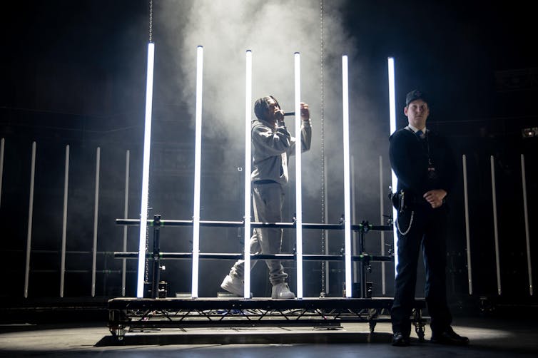 Digga D on stage behind white neon lights which look like a prison cell