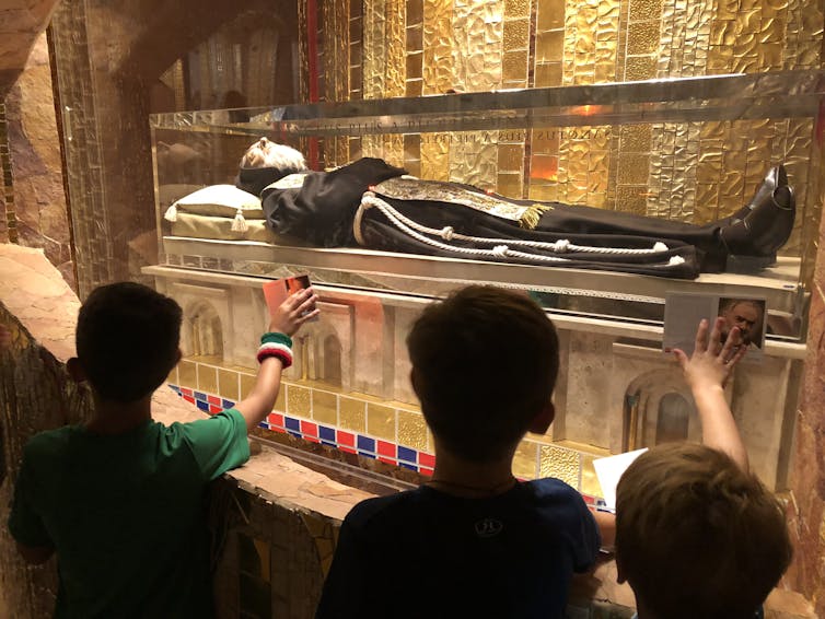 A saint's body, dressed in a black robe, lies inside a glass case and visitors extend their hands to touch it.