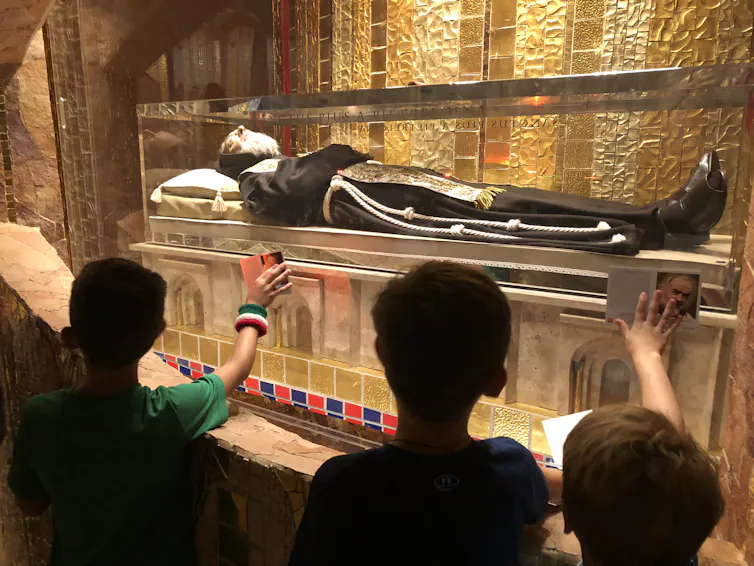 A saint's body, dressed in a black robe, lies inside a glass case and visitors extend their hands to touch it.
