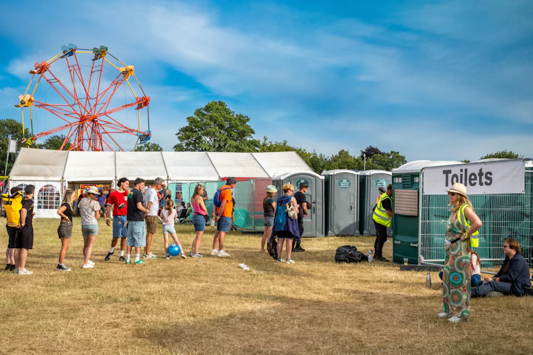 summer festival site with ferris wheeln, tents and a queue of people in summer clothes waiting by portaloo toilets on dry grass, blue sky