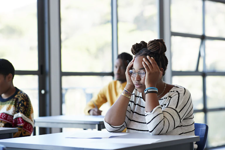 A female college student sits at a desk looking stressed with her hands against the sides of her face.