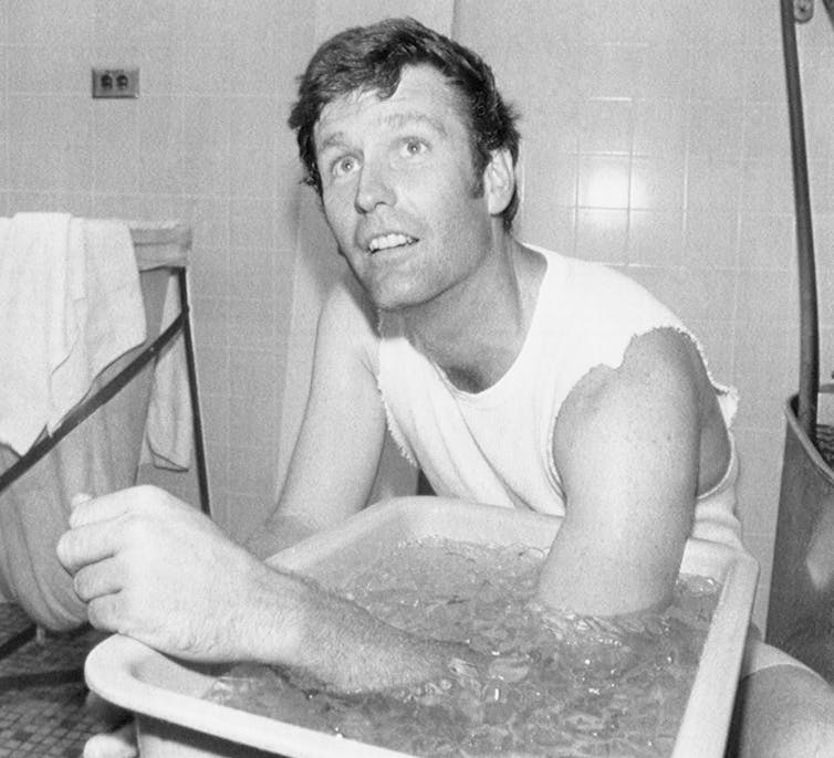 A man in a white cutoff T-shirt chills his elbow in a container of ice water.