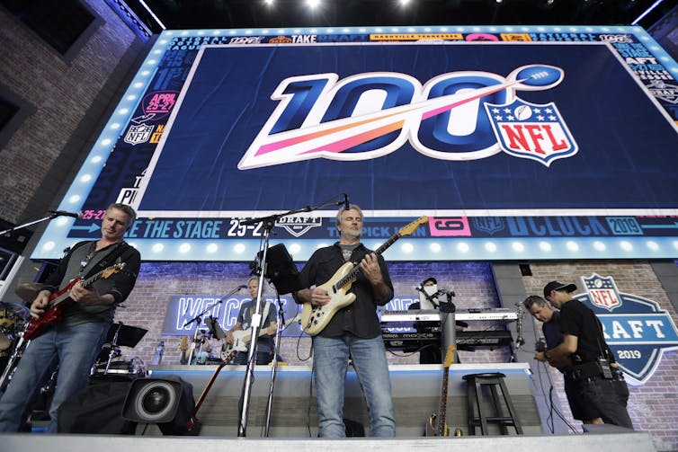 A band plays on a stage in front of a lit banner with the number 100 on it.