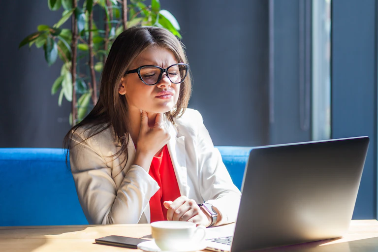 A young woman in a blazer holds her throat in pain while looking at a laptop.