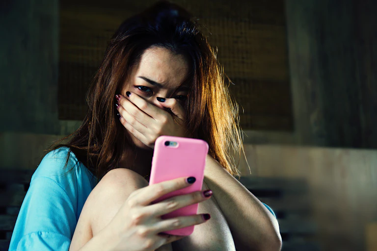 Young woman looking at her pink smartphone with a shocked expression, hand over mouth.