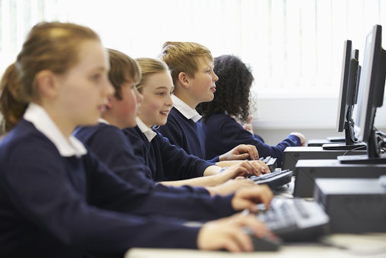 A side view of school students working in a row on desktop computers.