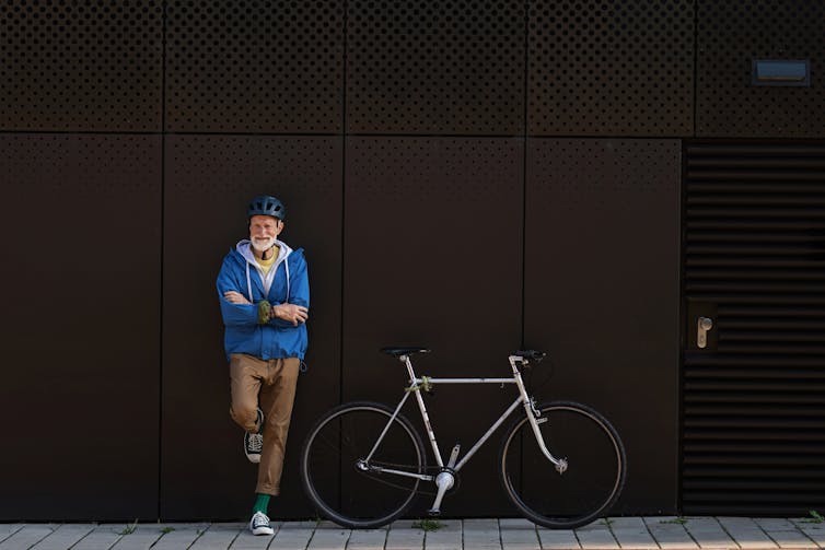 A tall older man leans against a wall while his bicycle is parked nearby.