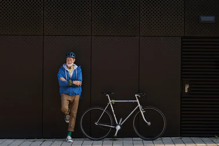 A tall older man leans against a wall while his bicycle is parked nearby.