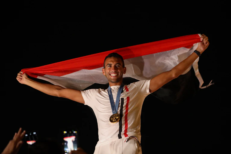 A man beams as he holds a red, white and black flag behind him, a gold medal around his neck.