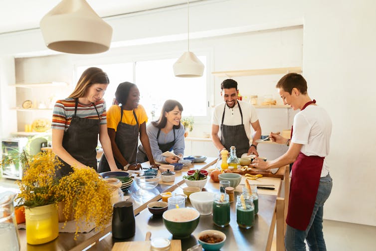 Five people wearing aprons around smart kitchen chopping and prepping food