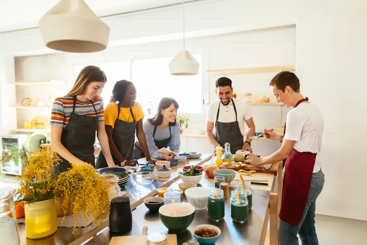 Five people wearing aprons around smart kitchen chopping and prepping food